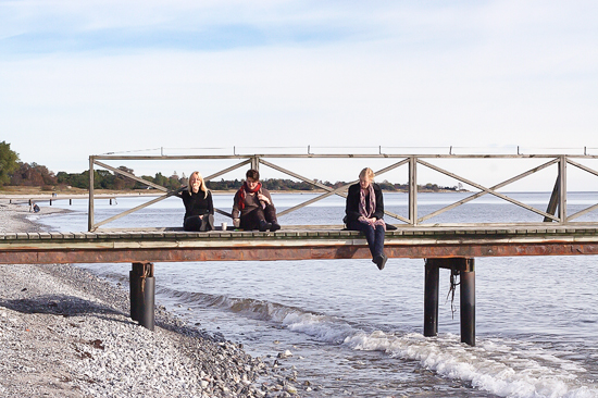 Three people are sitting on a pier at the shore.