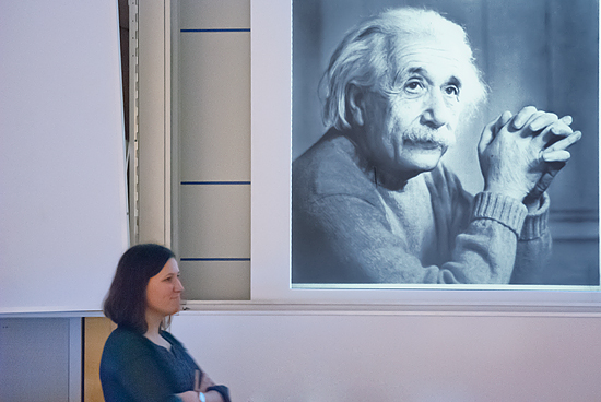 En person står i en föreläsningssal. I bakgrunden syns en presentation med en bild på Einstein.