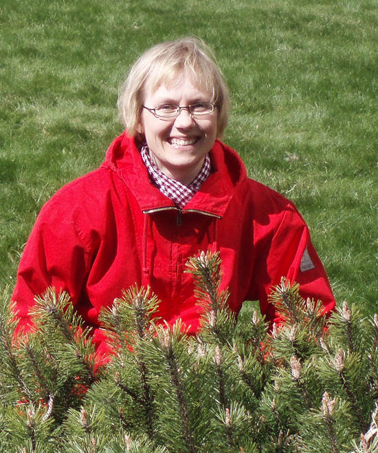 A smiling person is standing among some shrubbery. 