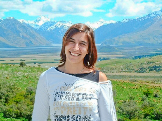 A smiling person stands in front of green grass and blue mountains.