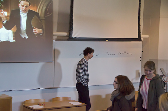 En person står längst fram i en föreläsningssal medan flera personer är på väg in.