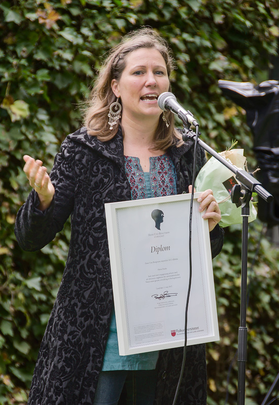 En person står utomhus och talar i en mikrofon. I handen håller hen en tavla med texten Diplom, resten syns inte.