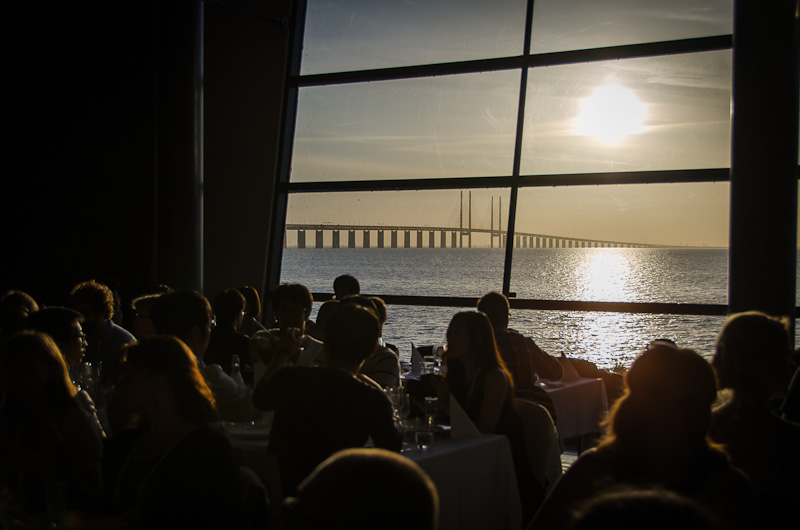 Genom ett fönster i bakgrunden syns Öresundsbron och en sol som håller på att gå ner. I förgrunden syns människor i siluett. De sitter vid matbord.