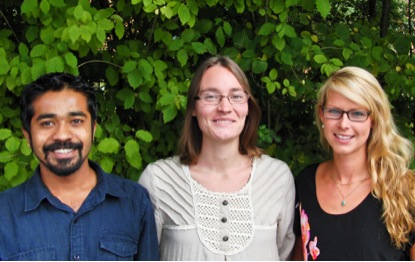 Three people are standing outside agains a green wall of leaves.