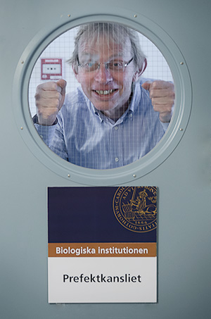 A person is looking out of a round window in a door. Beneath the window is a sign with the text Biologiska institutionen Prefektkansliet.