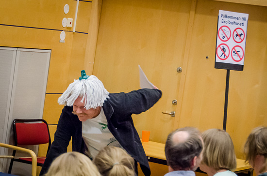 En person med en mopp på huvudet går framåtböjd  längst fram i en föreläsningssal.