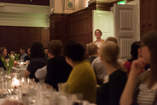 A lot of people are sitting at a dinner table. On person is standing up talking.