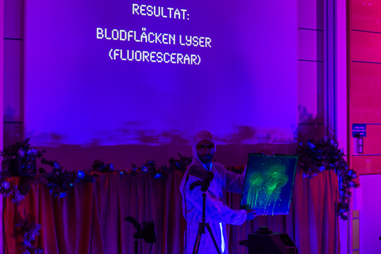 En föreläsningssal badande i blått ljus. I bakgrunden finns en projektorduk där det står "Resultat: blodfläcken lyser (fluorescerar)".