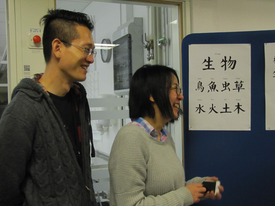 Two smiling people are standing with calligraphy signs in the background.