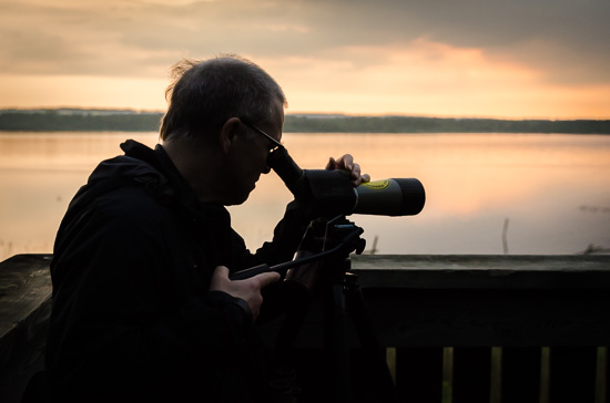 En person sedd i siluett tittar i en tubkikare. I bakgrunden är en sjö som färgas av den uppgående solen.