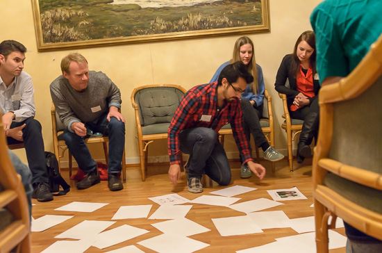 One person is sitting on the floor with a lot of papers in front of him. Other people are looking from behind.