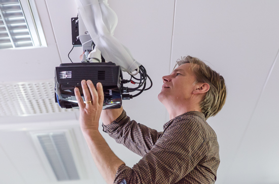 A person is standing looking at a video projector in the ceiling. 