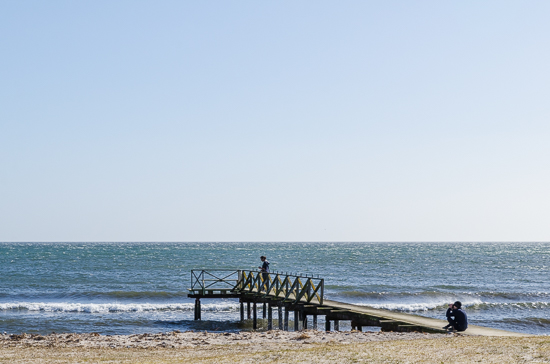 A person is standing alone on a pier. Another person is sitting taking photos.