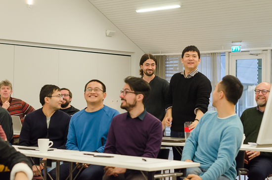 Some smiling persons are standing and sitting behind tables.