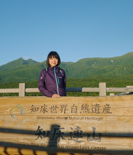 A person is standing behind a wood sign with Chinese signs and a text saying Shiretoko World Natural Heritage.